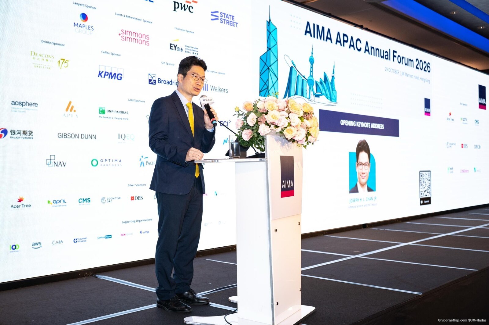 AIMA APAC Annual Forum 2026 - the Acting Secretary for Financial Services and the Treasury speaking at a podium in front of the forum's own LED wall at the JW Marriott Hotel Hong Kong