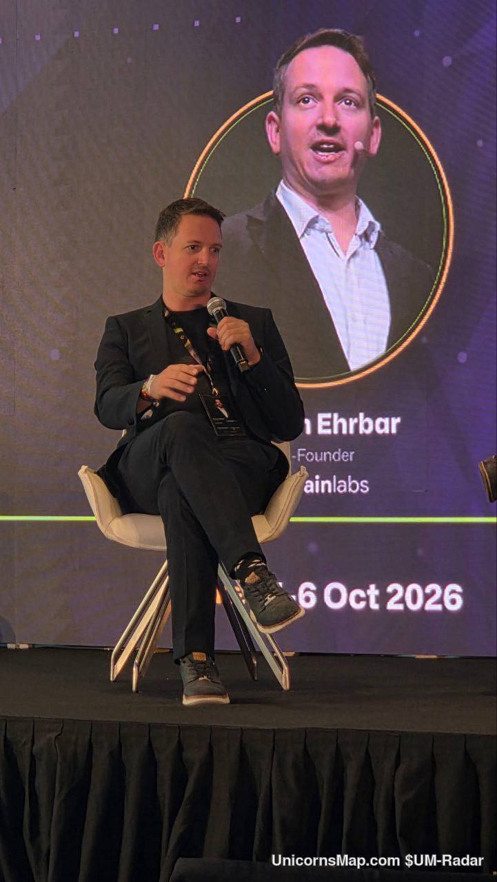CoinFerenceX The Best Event Singapore 2026 — a seated speaker with a handheld microphone on the Flower Field Hall stage at Gardens by the Bay, with the event's own large on-stage screen behind him carrying his name card (Ehrbar / Founder / Onchainlabs) and the on-screen date '6 Oct 2026'