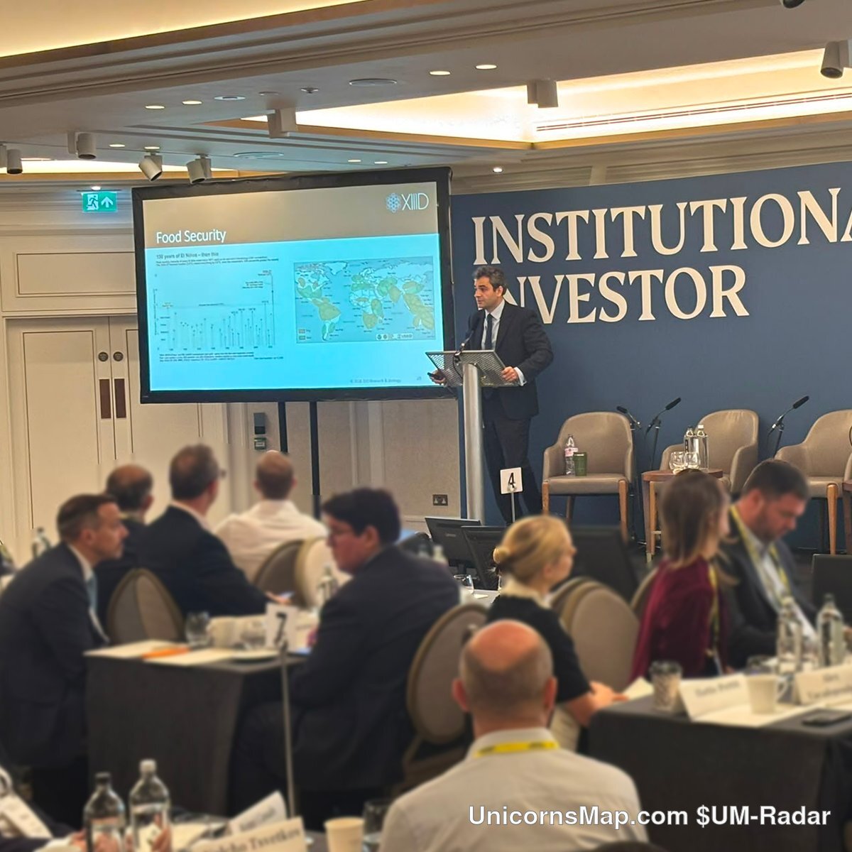 European Family Office Symposium 2026 — a keynote speaker at the lectern on the Institutional Investor stage in London, a presentation screen showing a Food Security slide with a world map, and the seated audience in the foreground