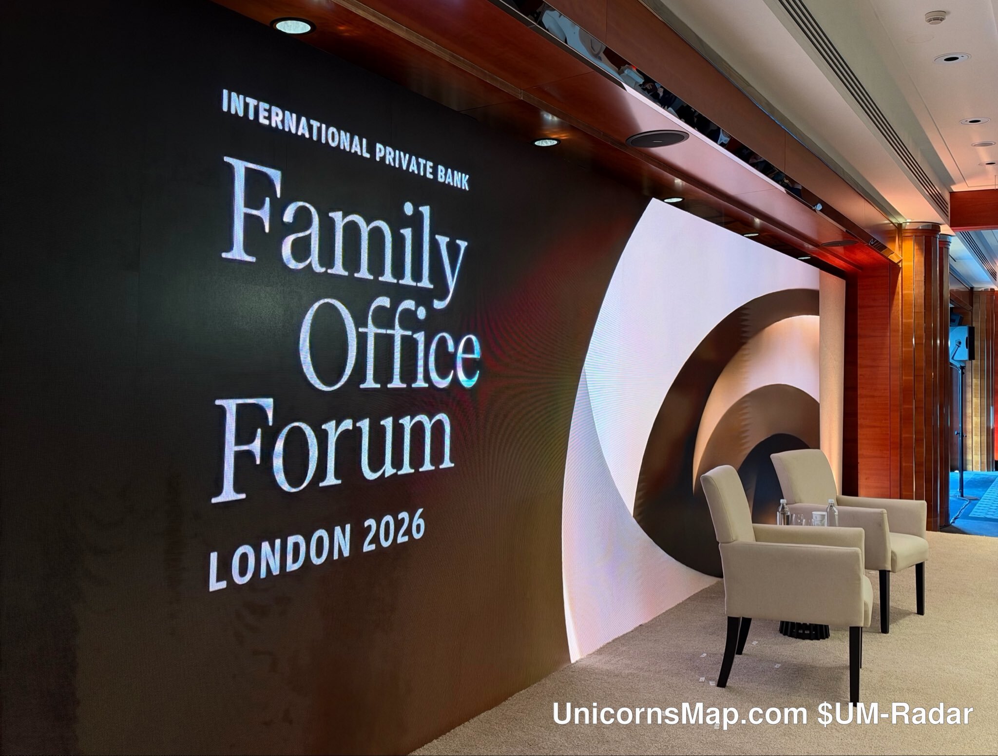 J.P. Morgan International Private Bank Family Office Forum London 2026 — the empty pre-session stage set: the organizer's own LED wall reading 'INTERNATIONAL PRIVATE BANK / Family Office Forum / LONDON 2026' beside two empty cream armchairs on a carpeted floor, with the venue's timber and glass interior behind; no people in frame