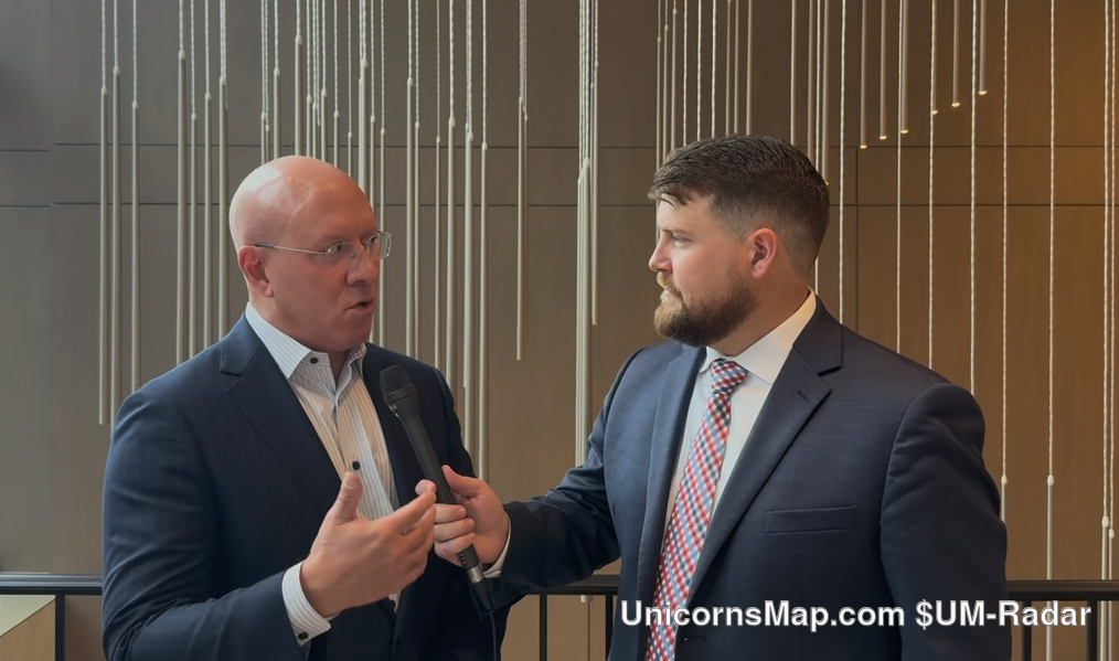 5th Annual Private Wealth Carolinas Forum 2026 — two suited men in an indoor hotel space with a hanging light-rod installation behind them: one bald, in glasses and a dark suit with an open-collar shirt, speaking and gesturing; the other, bearded, in a navy suit and patterned tie, holding a microphone towards him
