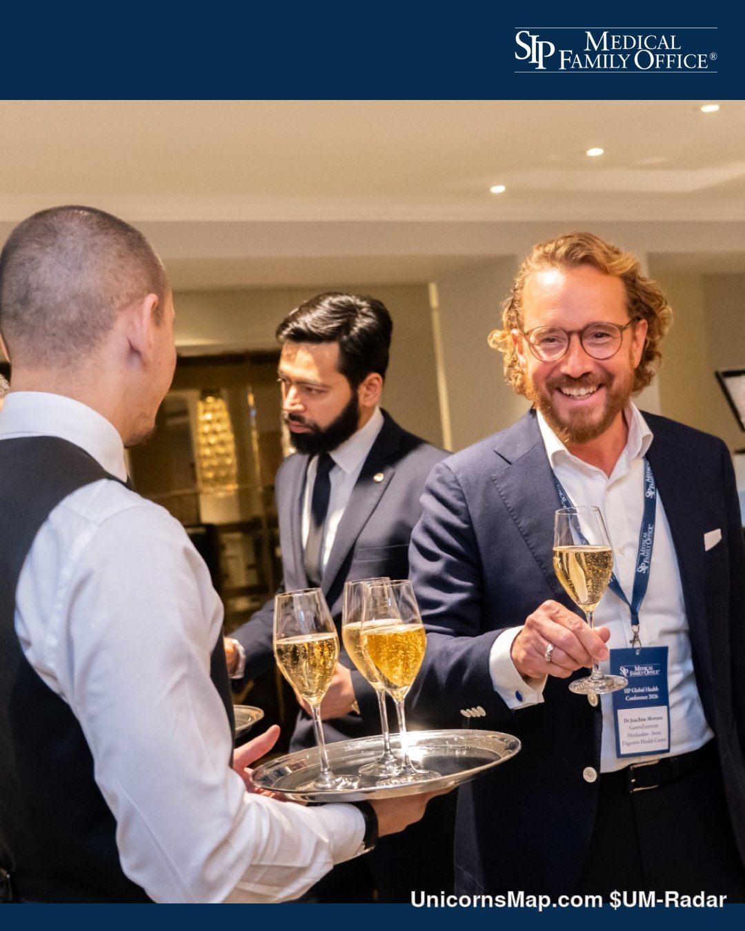 Networking reception at the SIP Global Health Conference 2026, The Langham, London: a waiter in a black waistcoat carrying a silver tray of champagne flutes to a smiling delegate in a navy blazer holding a glass, a second delegate behind them, delegate lanyards visible but unreadable