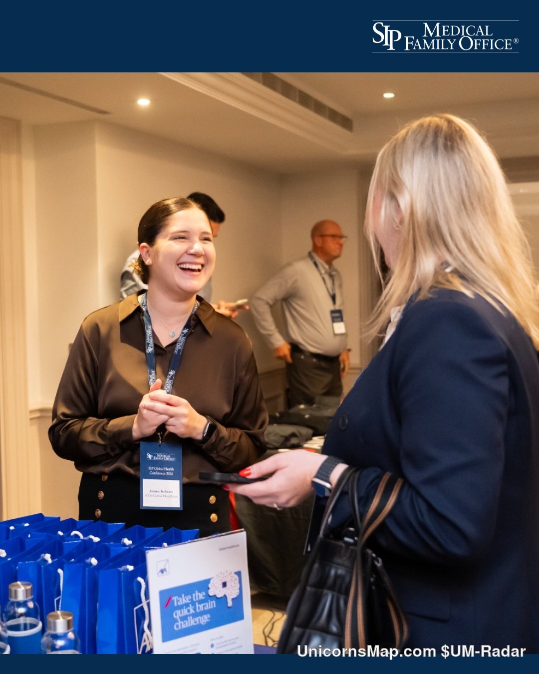 Registration and networking desk at the SIP Global Health Conference 2026, The Langham, London: two delegates talking beside a table of blue branded gift bags and water bottles, a 'Take the quick brain challenge' sign on the table, further delegates and a doorway behind