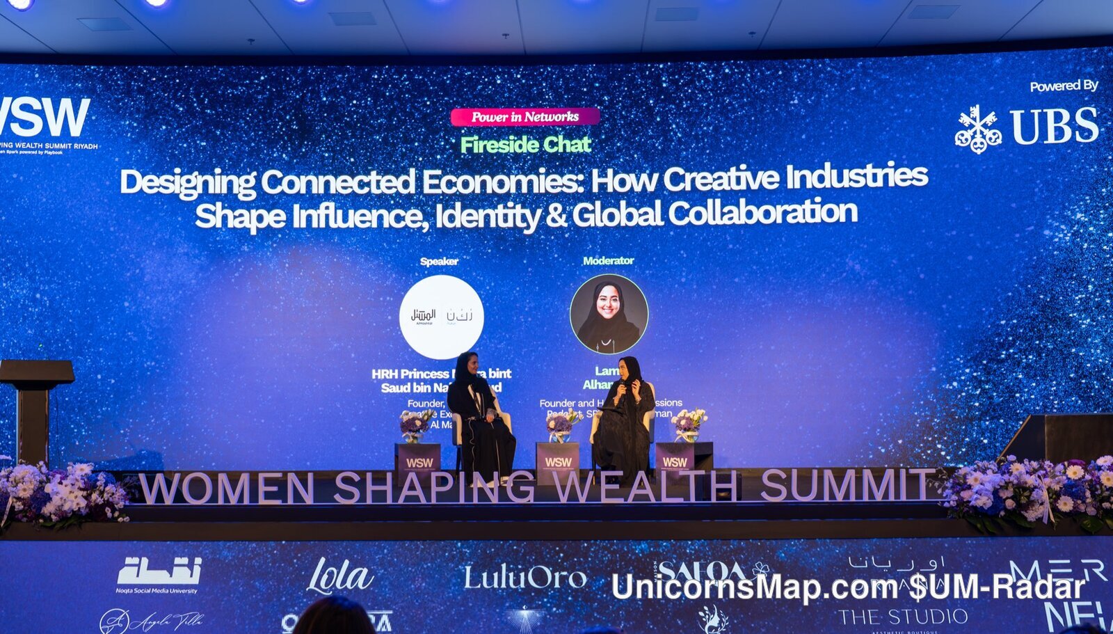 Women Shaping Wealth Summit 2026, Riyadh — wide day-of room frame: the event's own 'WOMEN SHAPING WEALTH SUMMIT' LED band and WSW stage screen carrying the 'Power in Networks — Fireside Chat' slide 'Designing Connected Economies: How Creative Industries Shape Influence, Identity & Global Collaboration' with the Speaker and Moderator name cards, the 'Powered By UBS' corner, the sponsor band and the audience seen from behind
